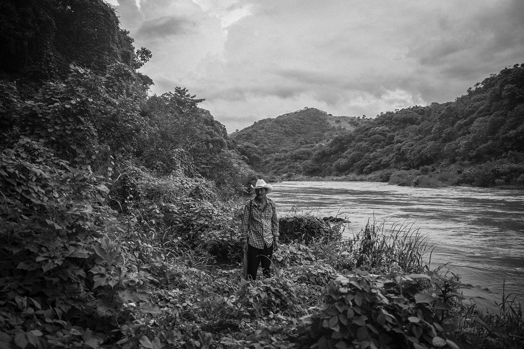 Gerardo Leiva on the banks of the Lempa River, at the site where he helped dozens of people cross on Mar. 18, 1981. He remembers that to get to this point, his “compas” had to fight off the army as people made their way toward Honduras.
